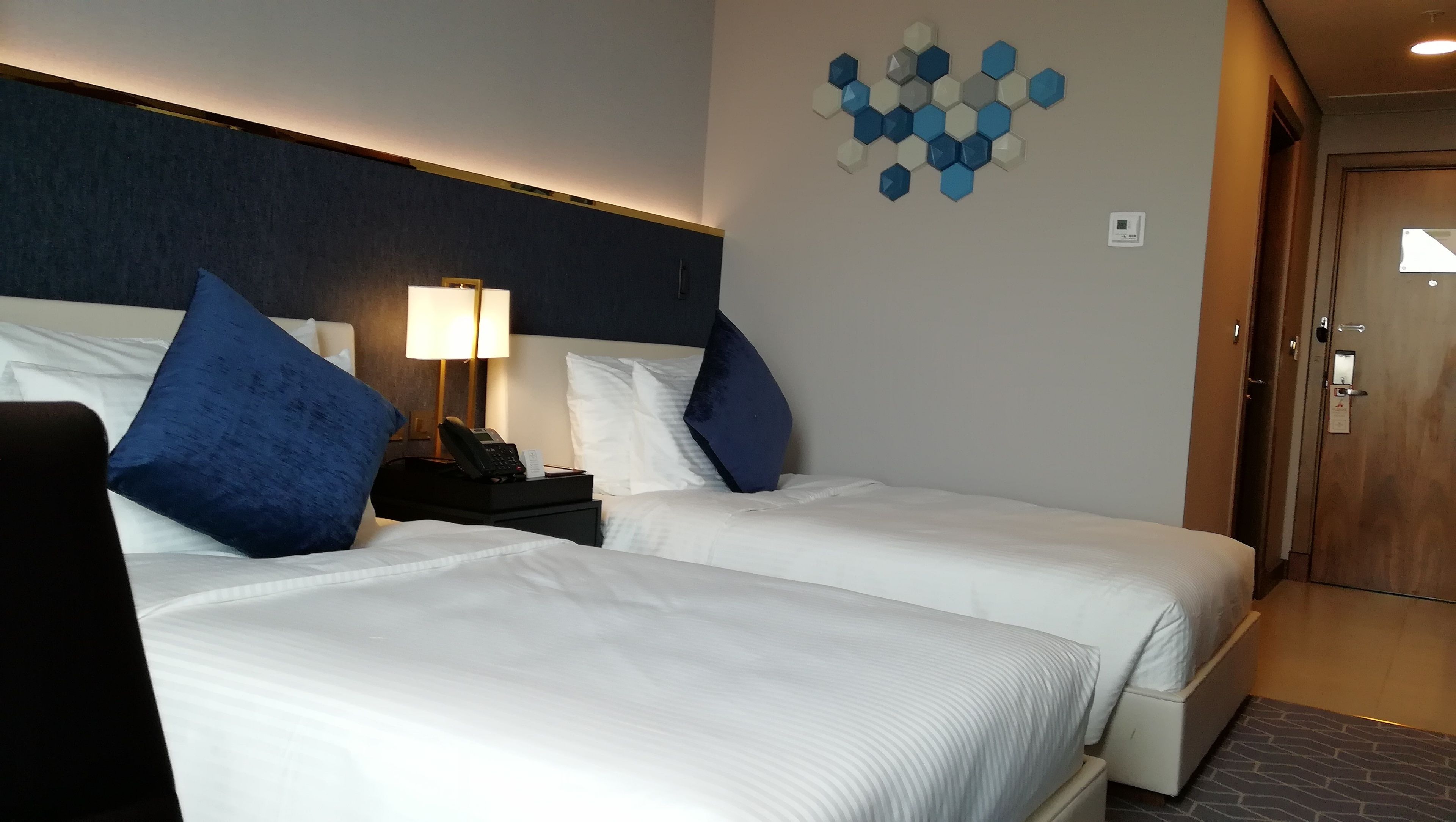 Quarto de hotel com duas camas, uma mesa de cabeceira com abajur, telefone e um ornamento na parede.