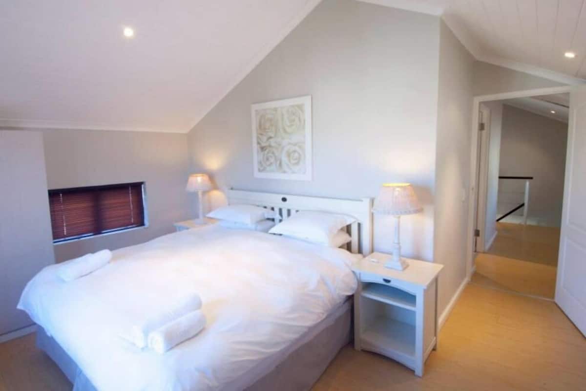 Une chambre à coucher avec un grand lit, des tables de chevet, une fenêtre avec des stores et une porte donnant sur une autre pièce.