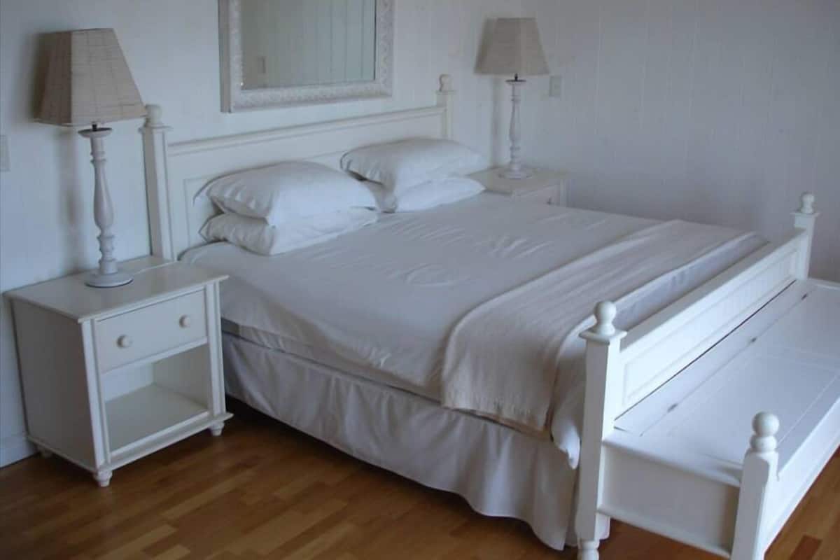 Un lit à baldaquin blanc, avec une table de chevet et une lampe, un miroir au-dessus du lit et un plancher en bois.