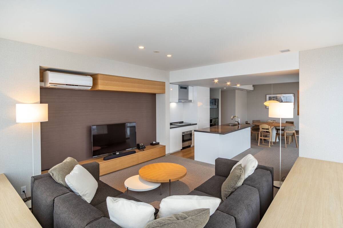 A modern living room with a sofa, a coffee table, a TV, and a kitchen area in the background.
