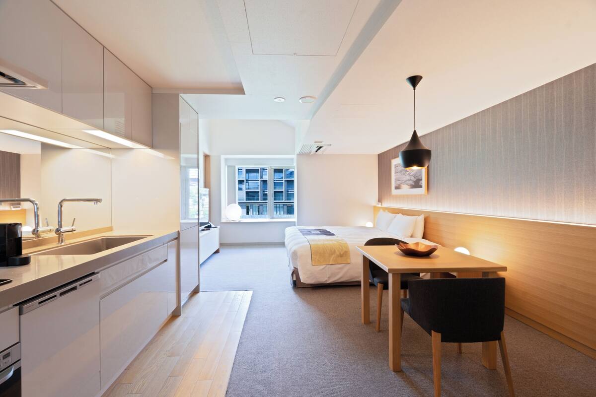 A modern hotel room with a kitchen, a dining area, and a bed.