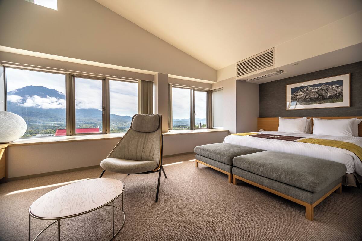 A modern hotel room with a large bed, a sofa, a chair, and a small table. There are large windows with a mountain view.