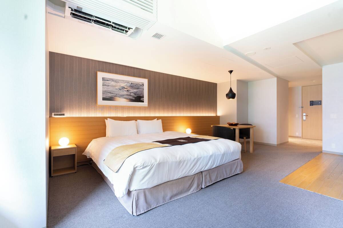 A modern hotel room with a large bed, bedside lamps, a wooden headboard, a framed picture, and a small table with a chair.