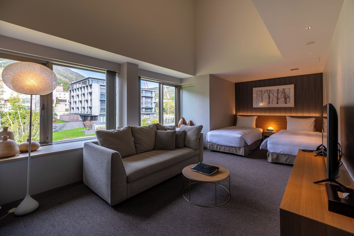 A modern hotel room with a sofa, two beds, a desk, and a large window offering a view of buildings and greenery.