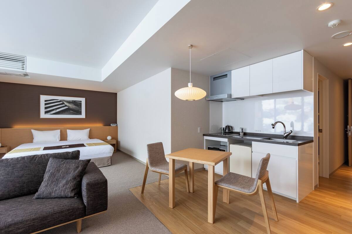 A modern hotel room with a bed, a sofa, a dining table with chairs, a kitchen area, and a pendant light.