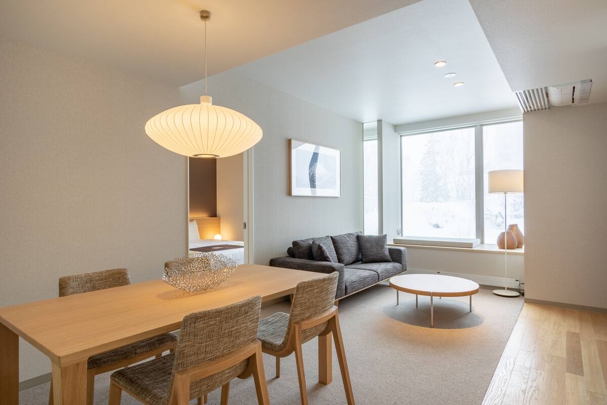 A modern living room with a dining area, featuring a wooden table, wicker chairs, a grey sofa, and a round coffee table.