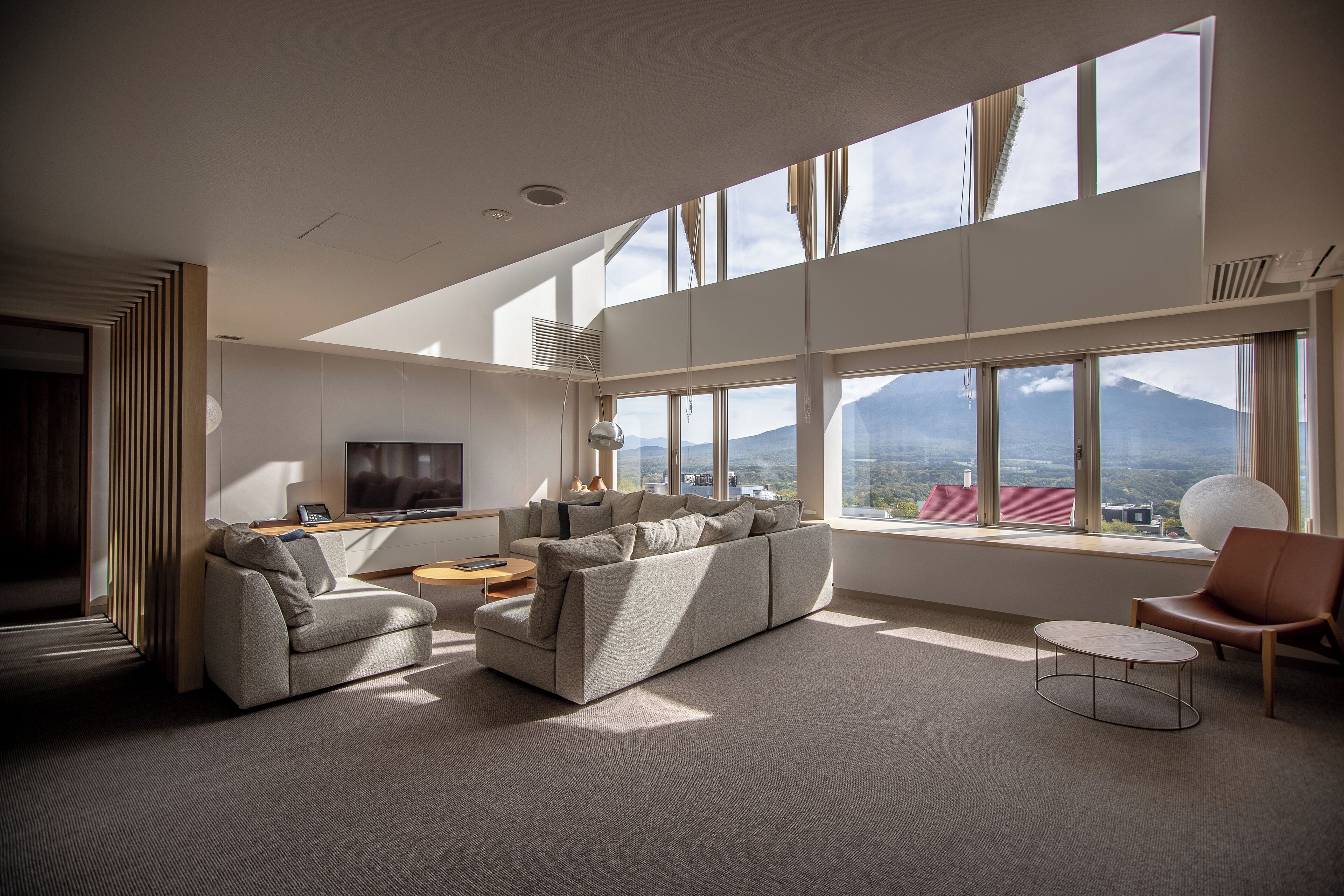 A modern living room with a sectional sofa, a coffee table, and a TV. Large windows offer a view of a mountain.