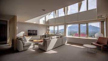 A modern living room with a sectional sofa, a coffee table, and a TV. Large windows offer a view of a mountain.