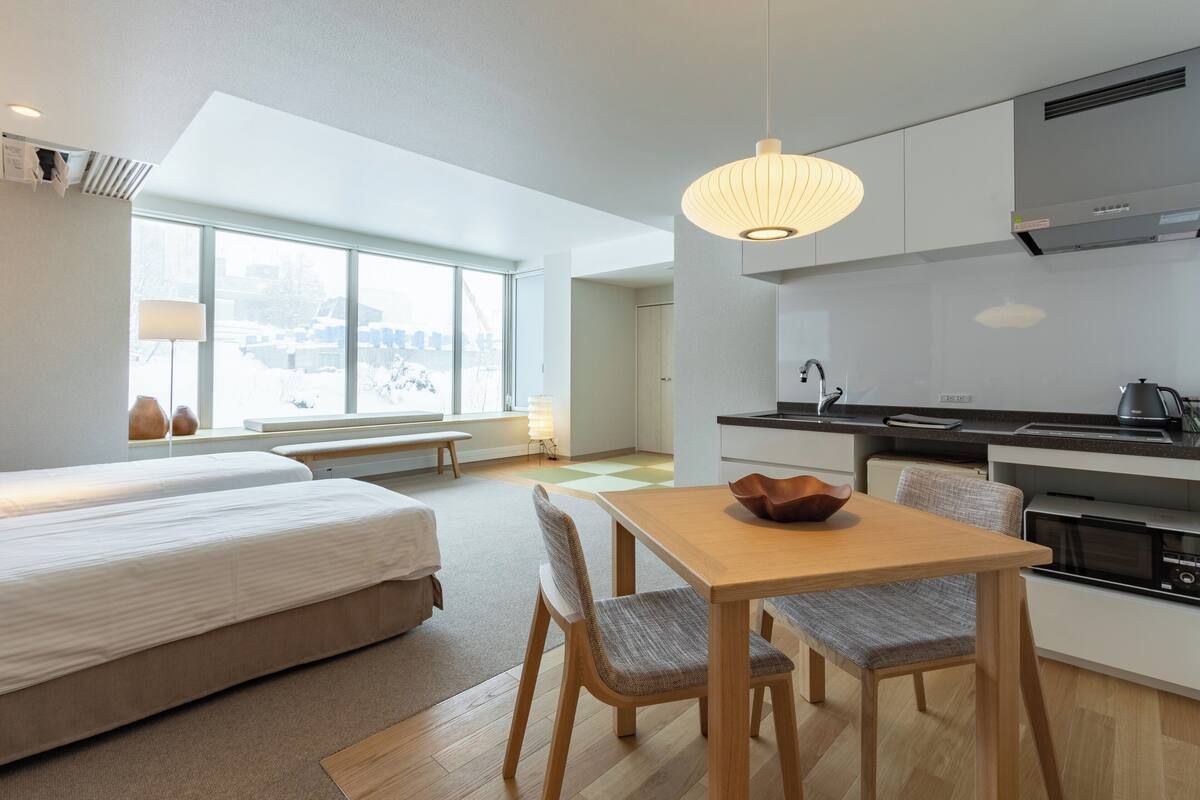 A modern hotel room with a bed, a dining area, a kitchen, and large windows.