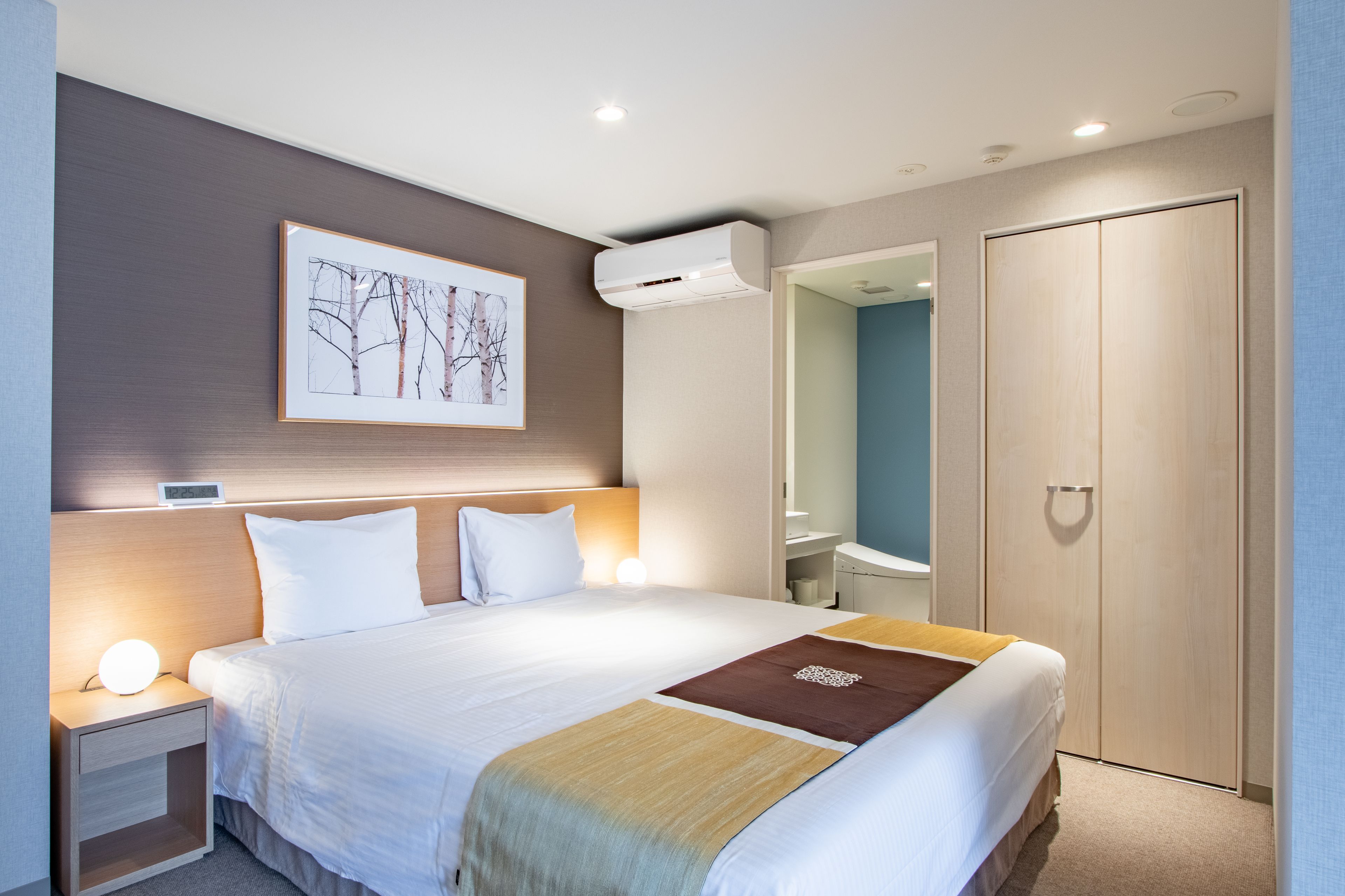 A hotel room with a bed, bedside tables, a wall-mounted air conditioner, a wardrobe, and a framed picture on the wall.