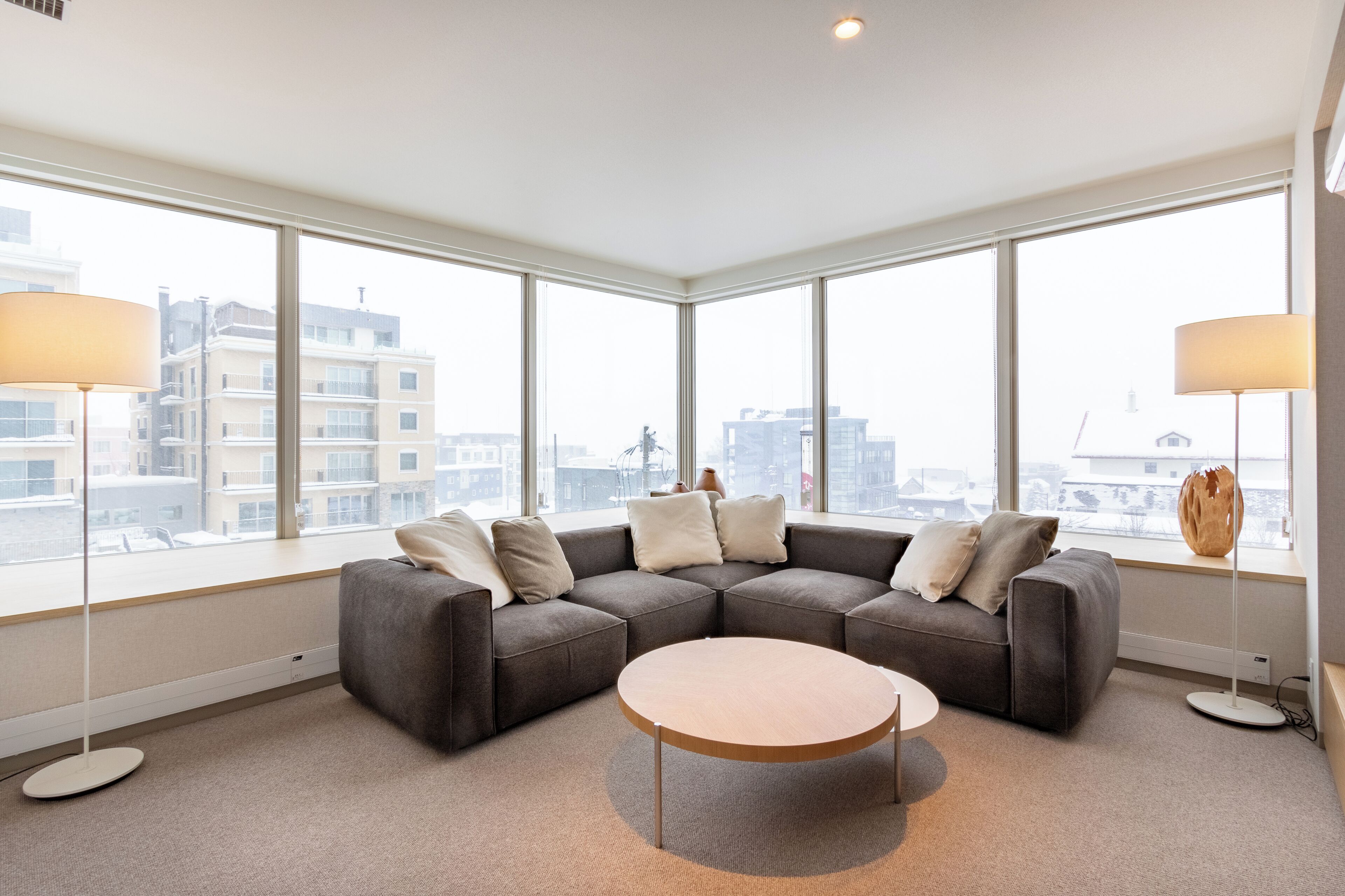 Una sala de estar moderna con un sofá seccional, una mesa redonda de centro y lámparas de pie. Amplias ventanas ofrecen vistas del paisaje urbano.