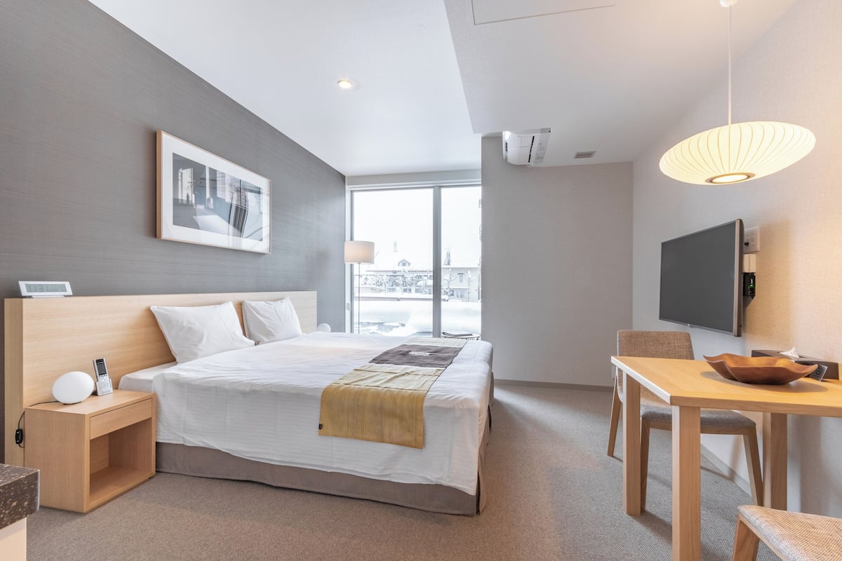 A modern hotel room with a large bed, a desk, a chair, a television, and a window with a view.