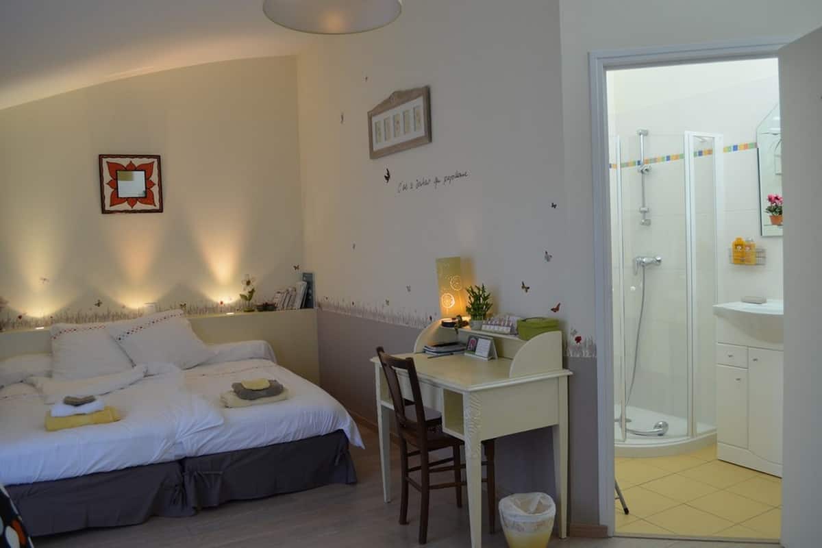 Quarto com cama, mesa de cabeceira, cadeira e pequena escrivaninha com luminária. É visível uma área de chuveiro por uma porta aberta.