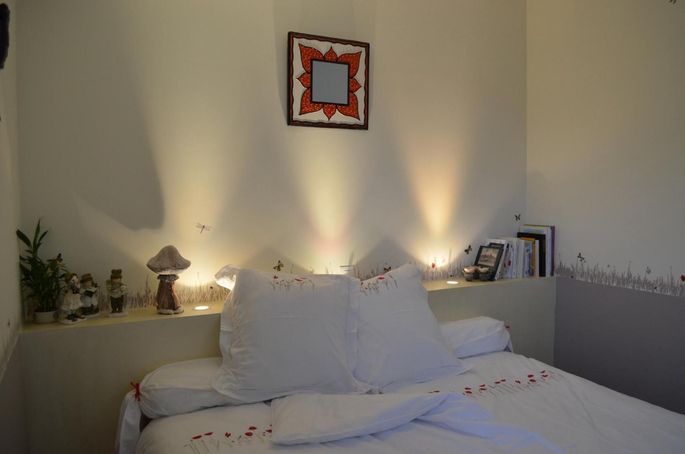 A neatly made bed with white bedding and pillows, a bedside table with a lamp, and a small shelf with decorative items.