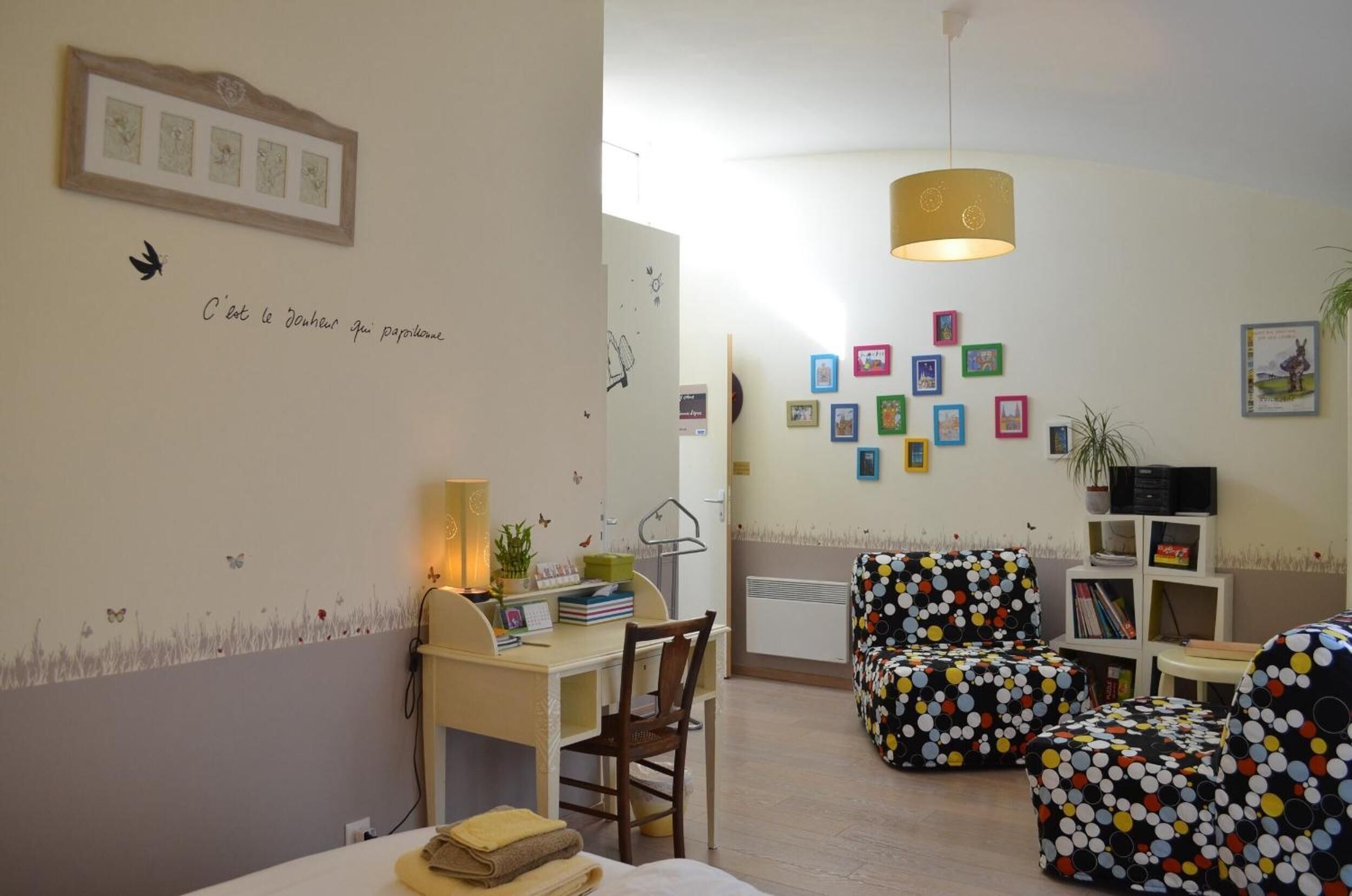 A cozy room with a desk, chair, and ottoman, a wall with framed pictures, and a ceiling light.