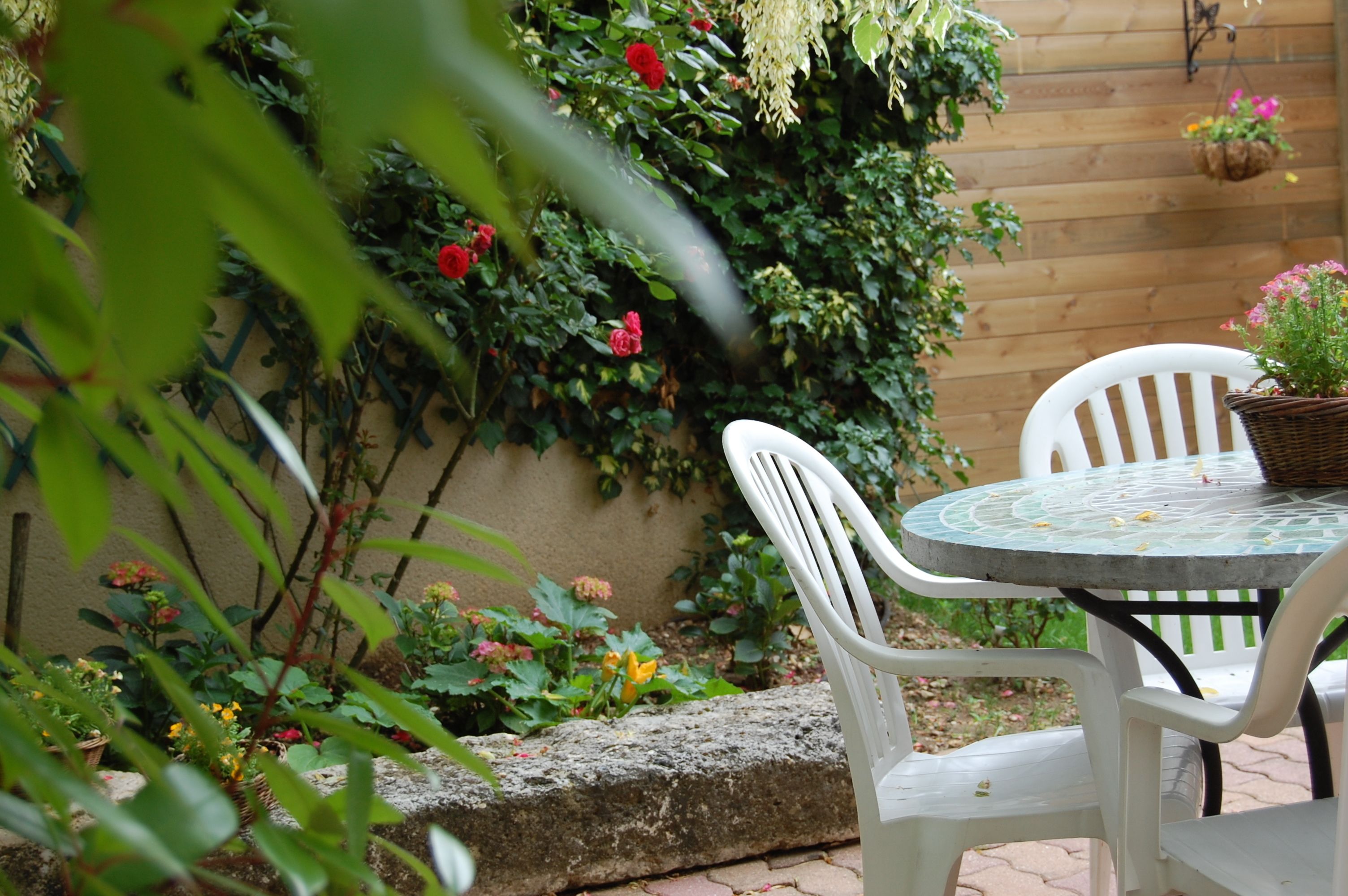 A patio with white plastic chairs and a round table, surrounded by a variety of plants and flowers.
