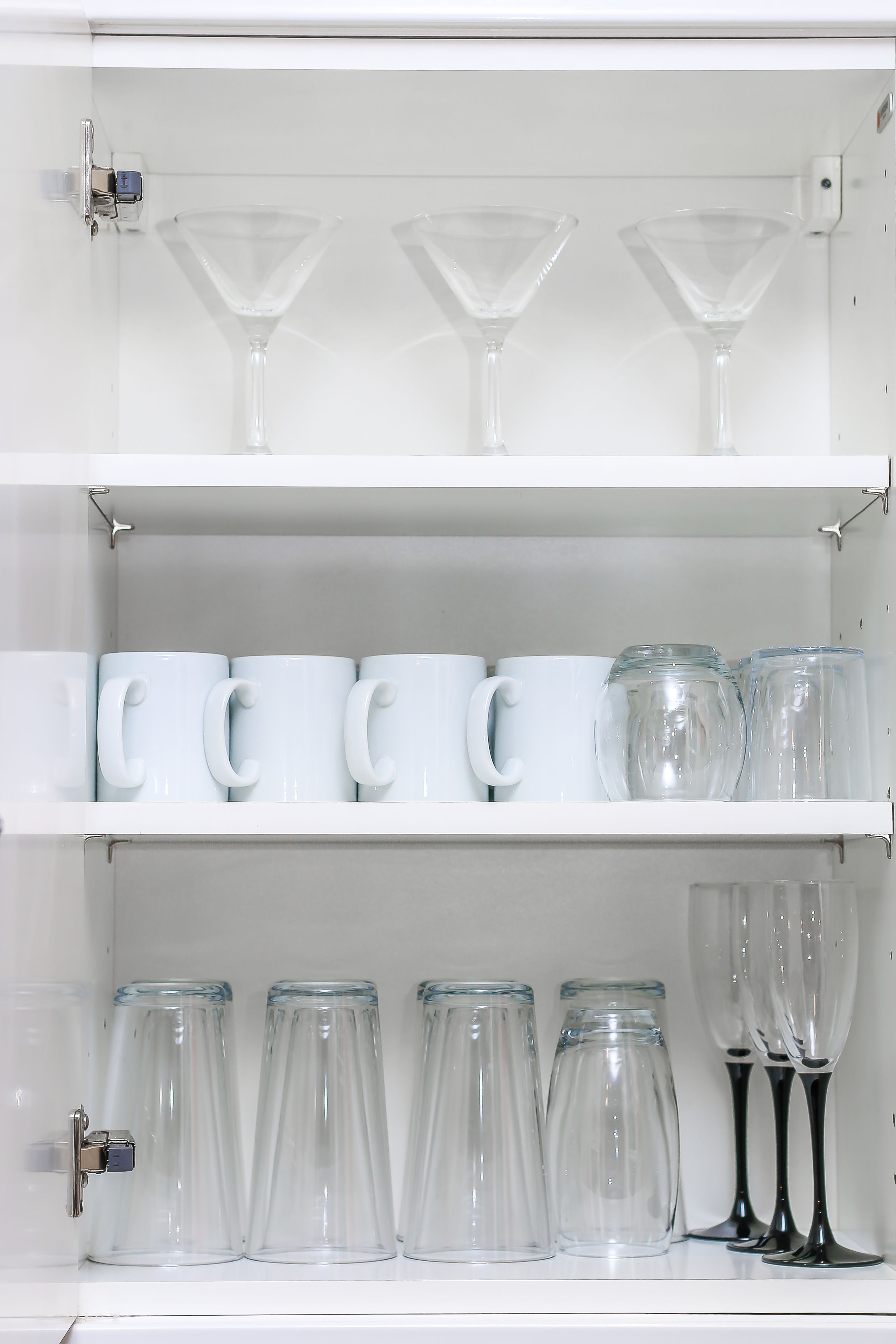 Een witte plankenwand met diverse glazen, waaronder heldere wijnglazen en witte mokken.
