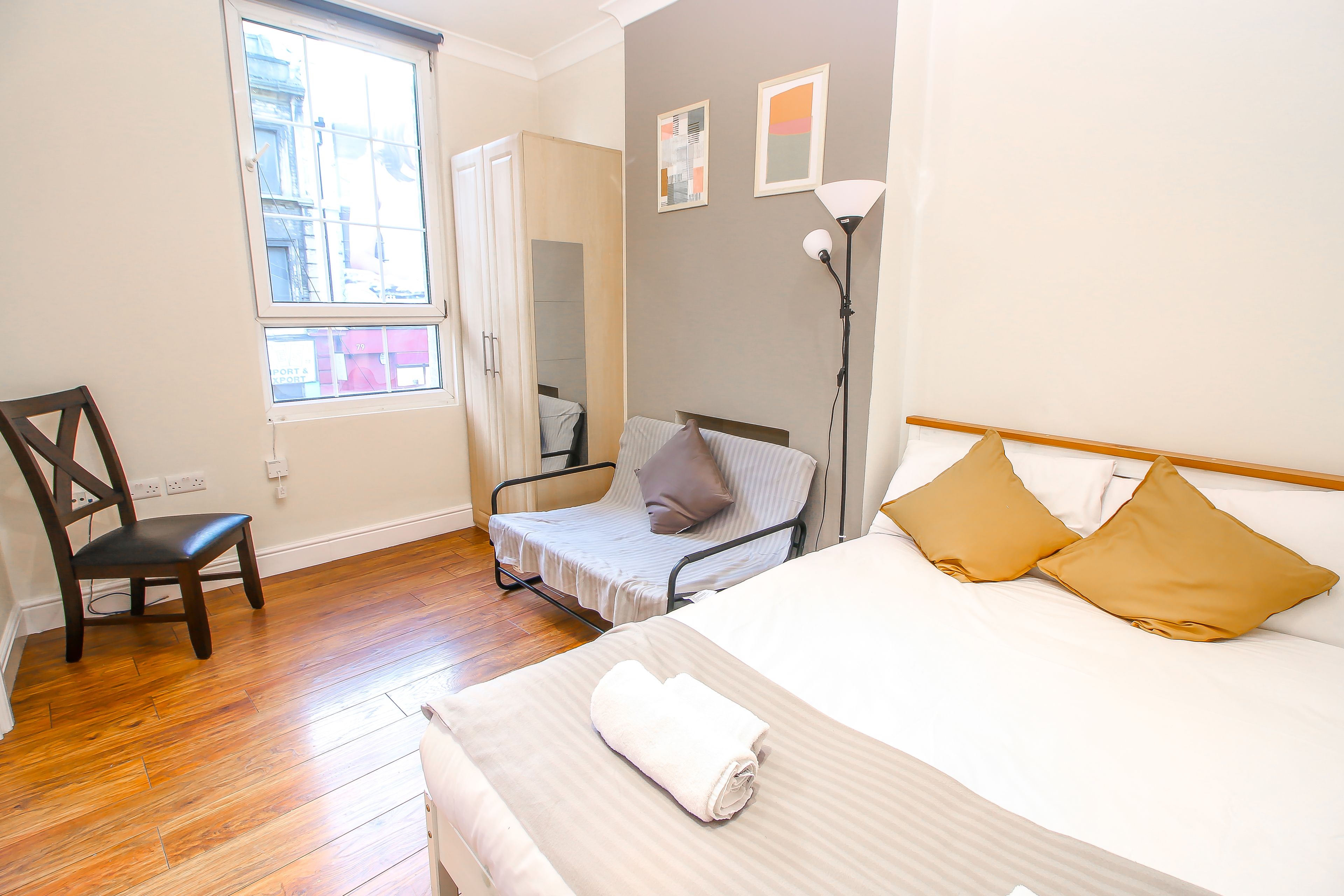 A hotel room with a wooden floor, a bed with two pillows, a chair, a bedside table, and a window with a view of buildings.