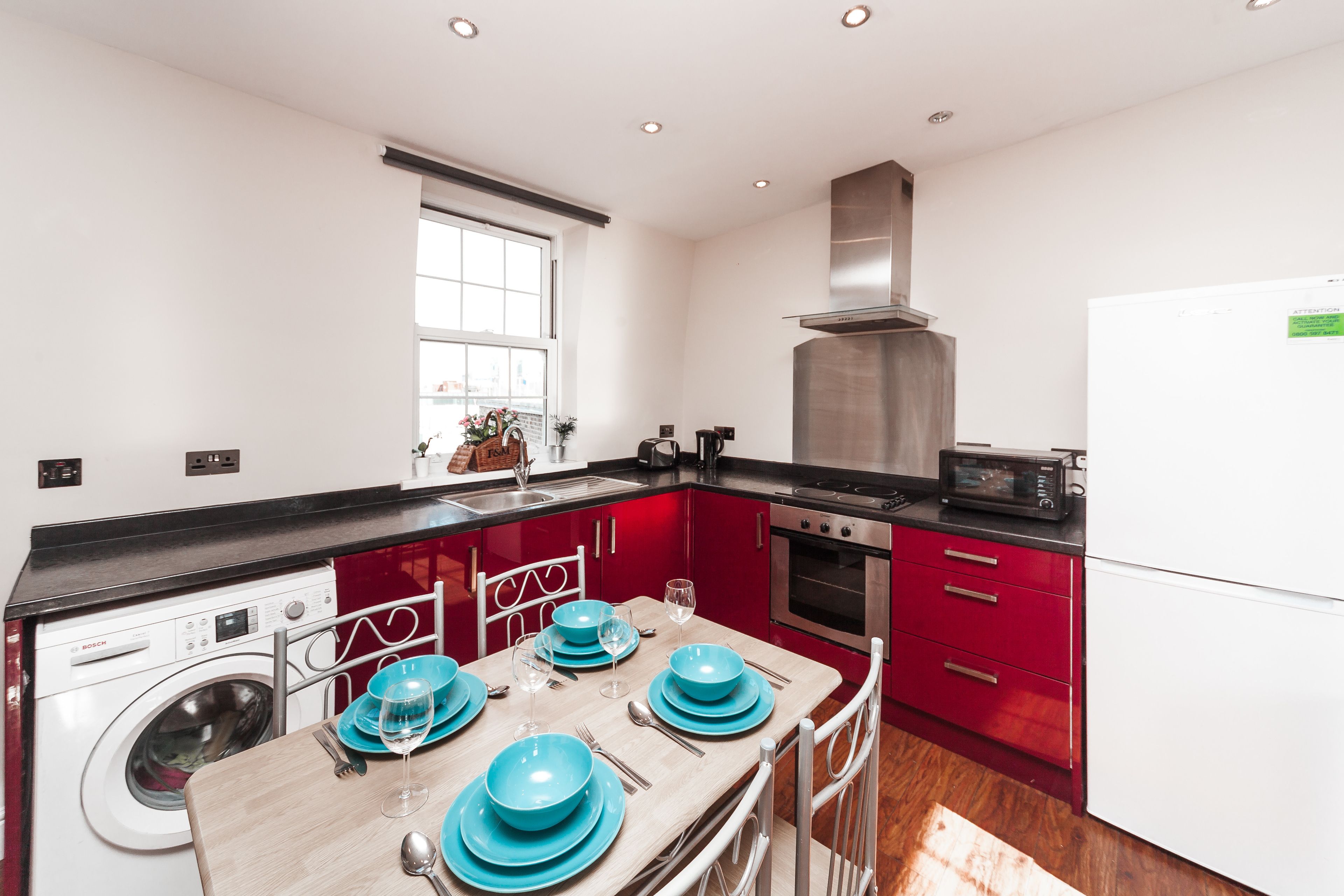A modern kitchen with red cabinets, a stainless steel range hood, a built-in oven, and a microwave. There is a washing machine and a sink with a window above it.