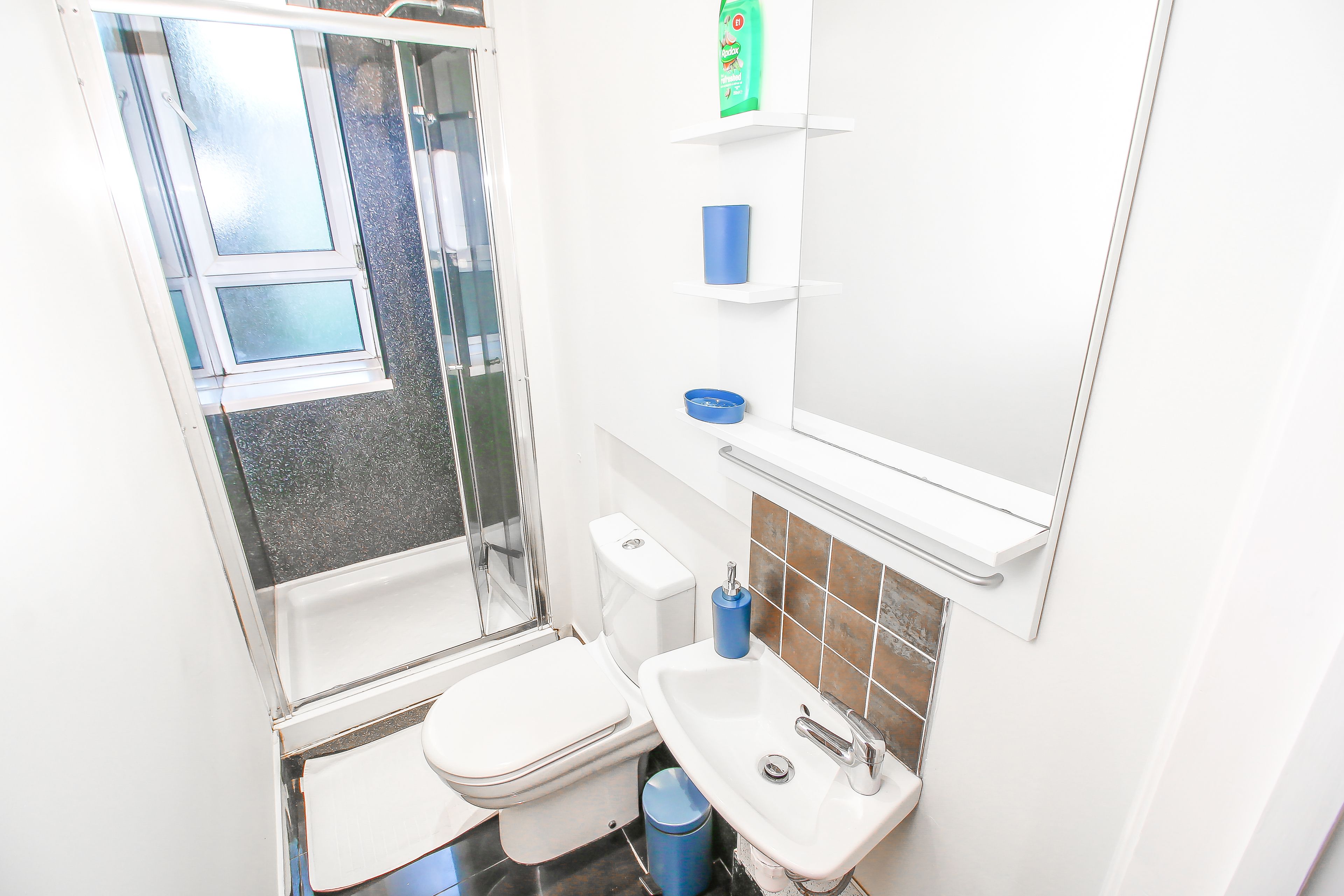 A small, white bathroom with a toilet, sink, and shower. There is a window and a mirror above the sink.