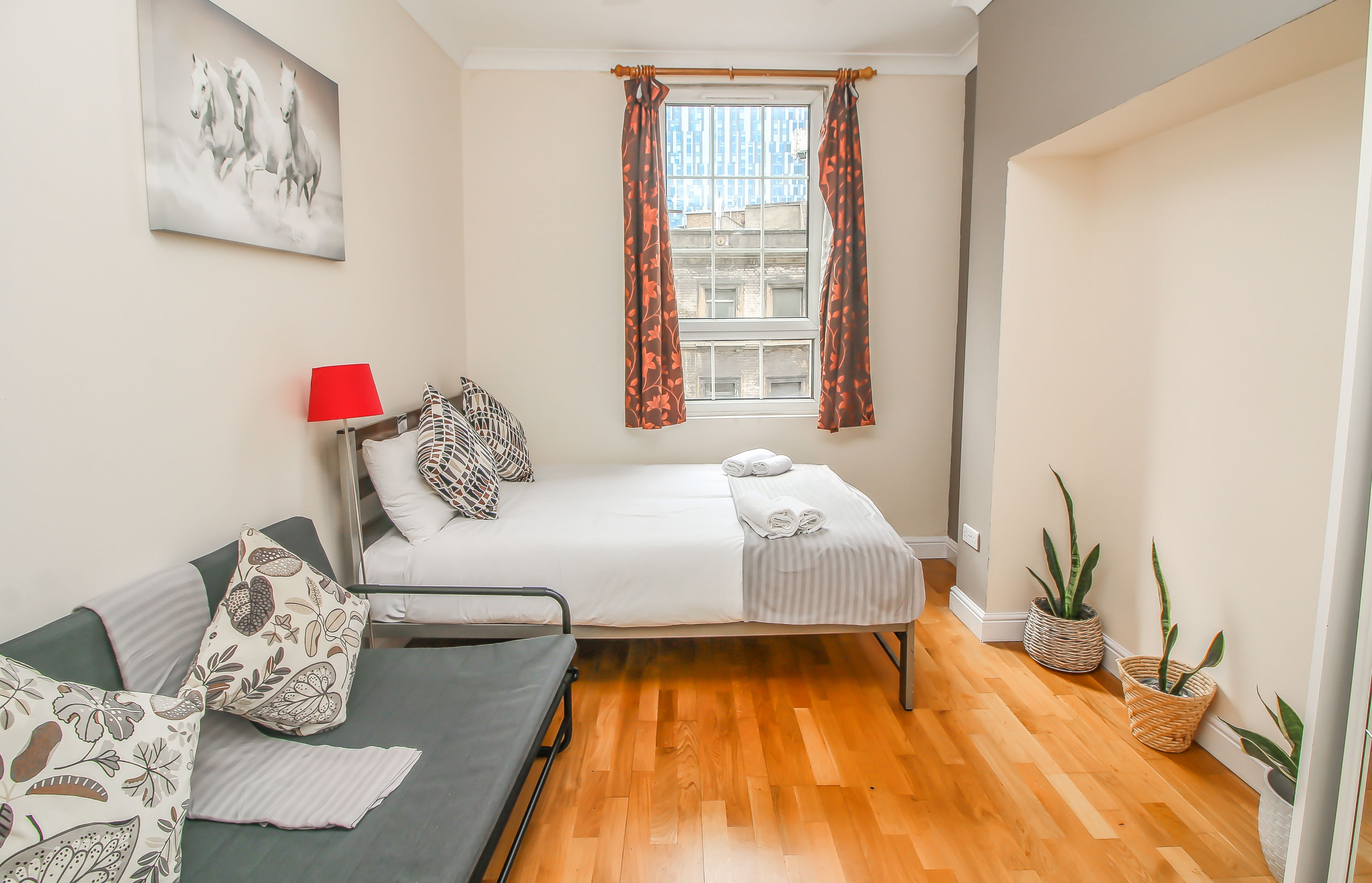 A room with a bed, a sofa, a red lamp, patterned cushions, a window with curtains, and potted plants.