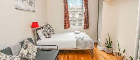 A room with a bed, a sofa, a red lamp, patterned cushions, a window with curtains, and potted plants.