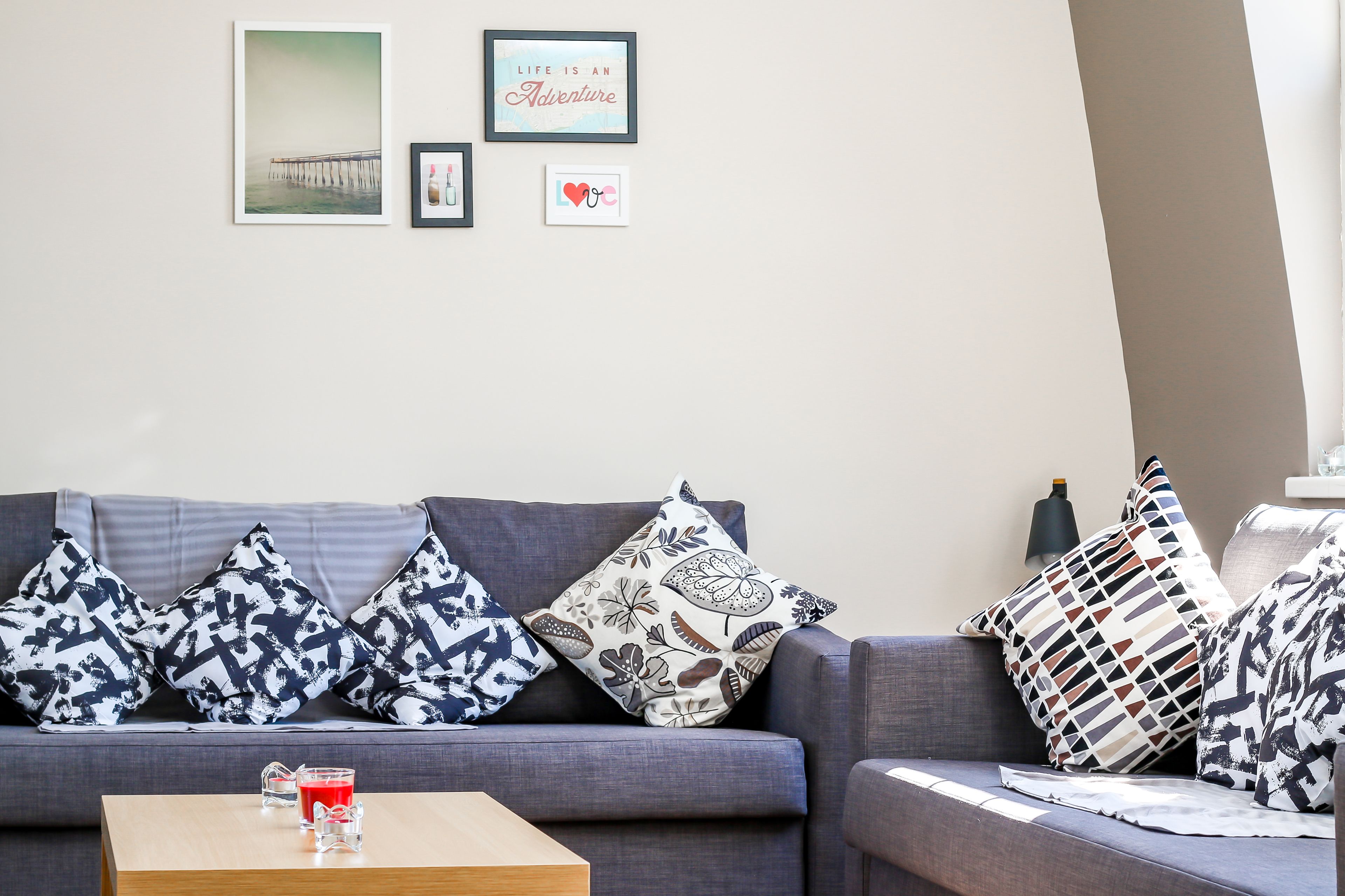 A living room with a grey sofa, patterned cushions, a wooden coffee table with a glass of red drink, and wall art including a quote and a heart symbol.
