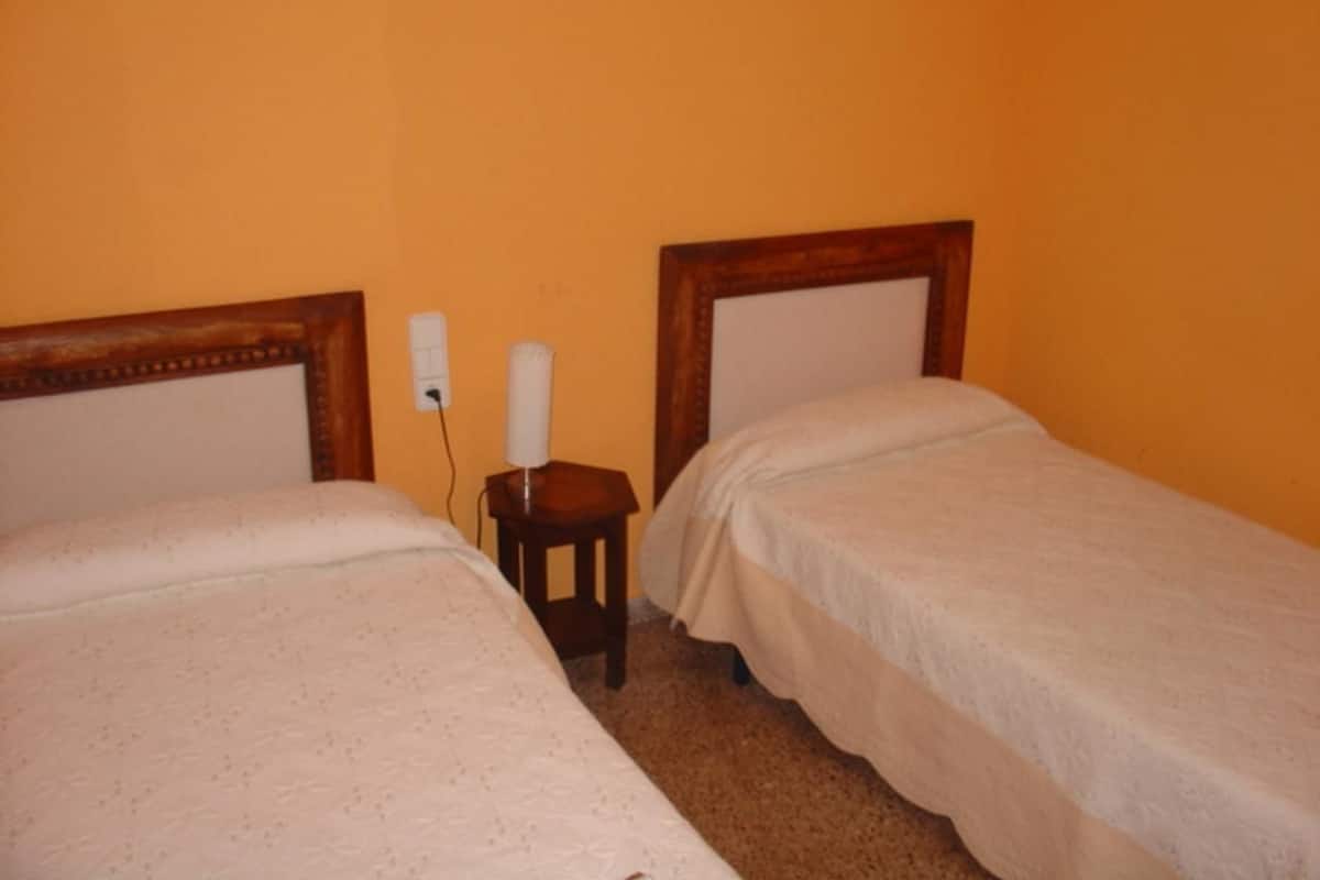 A room with two single beds, a small wooden nightstand with a lamp, and a plain orange wall.