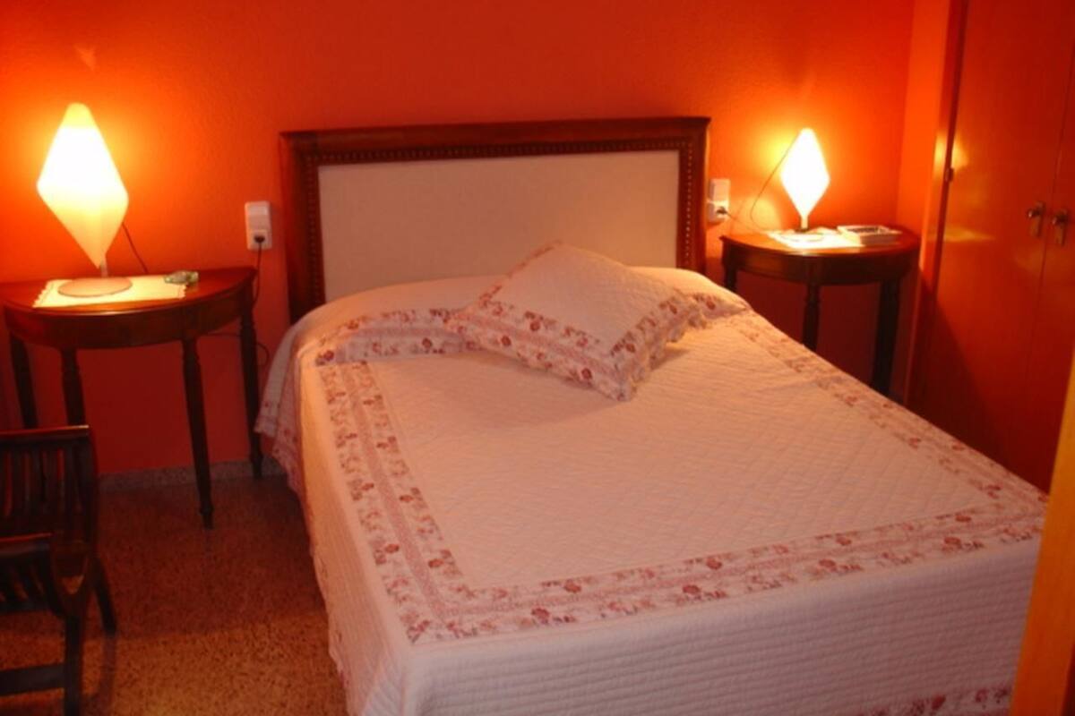 A neatly made bed with a floral-patterned bedspread and pillows, flanked by two bedside tables with lamps, in a room with orange walls.