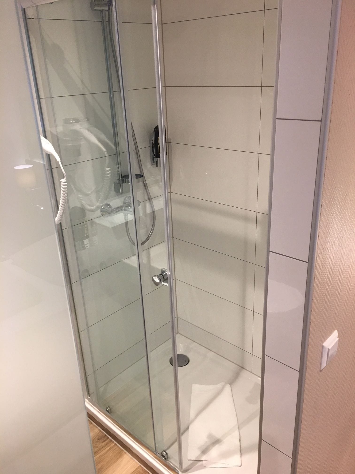 Une cabine de douche fermée par des parois en verre, dotée d’un pommeau de douche à main, d’un panneau de commande fixé au mur et d’un drain au sol.