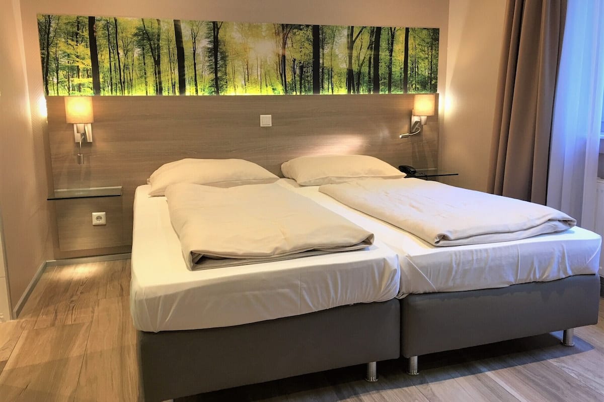 Una cama doble con dos almohadas, una mesita de noche, una lámpara de pared y un cuadro de un bosque.