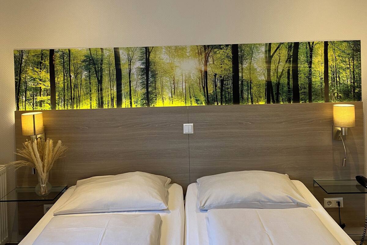 Un dormitorio con dos camas, un cuadro colgado en la pared que muestra un bosque, y lámparas de noche.
