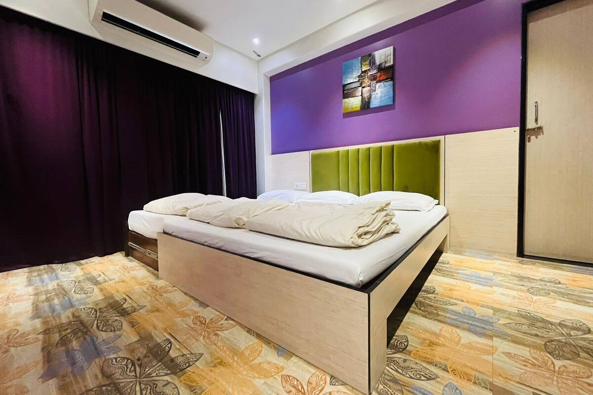 Una habitación de hotel moderna con cama, aire acondicionado, paredes moradas, cabecera verde y alfombra estampada.