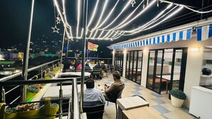 Un área de comedor al aire libre con mesas y sillas, vista de la ciudad de noche, y luces colgantes.