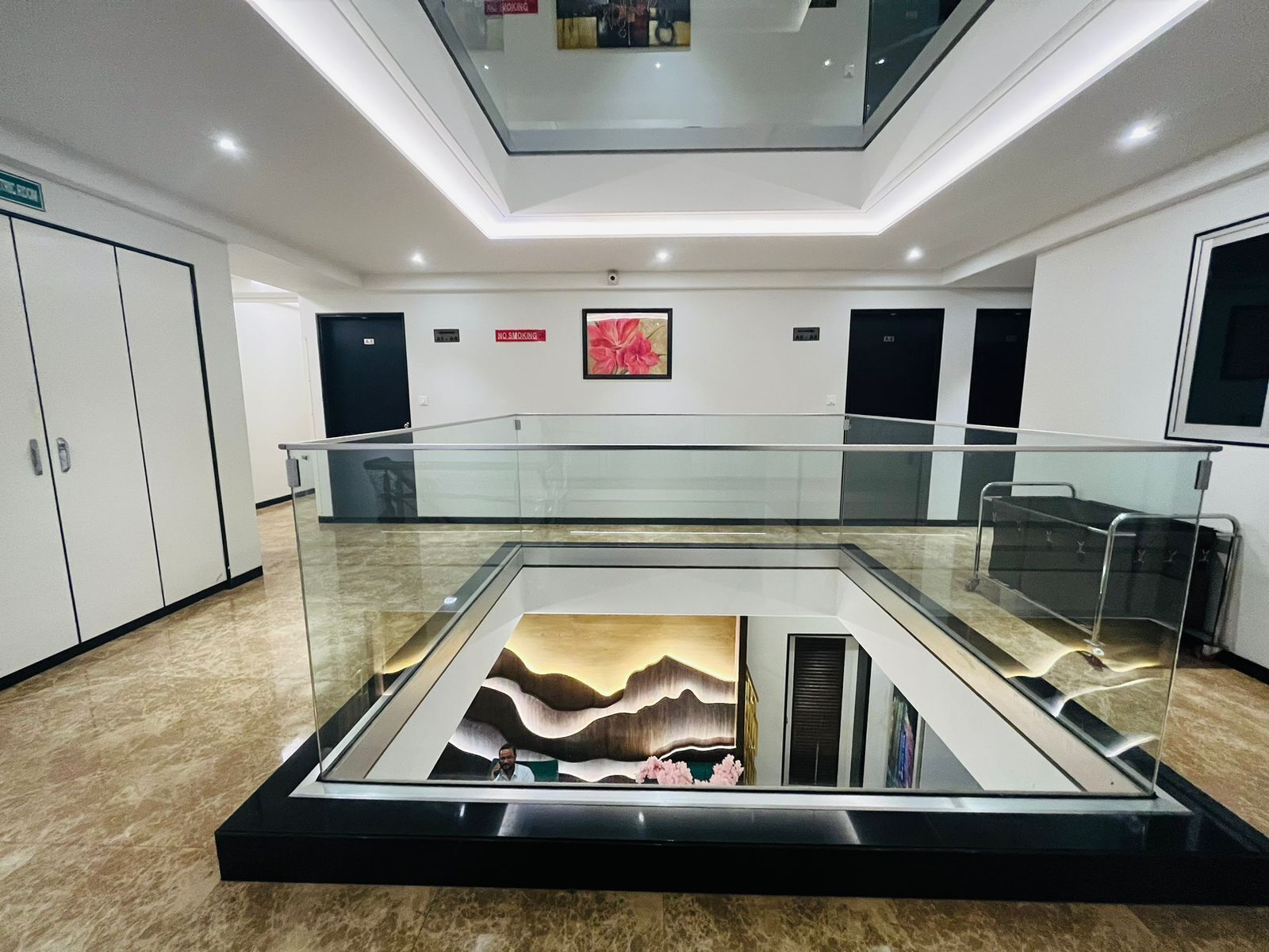A modern hotel lobby with a glass-bottomed atrium, a no-smoking sign, and a large painting on the wall.