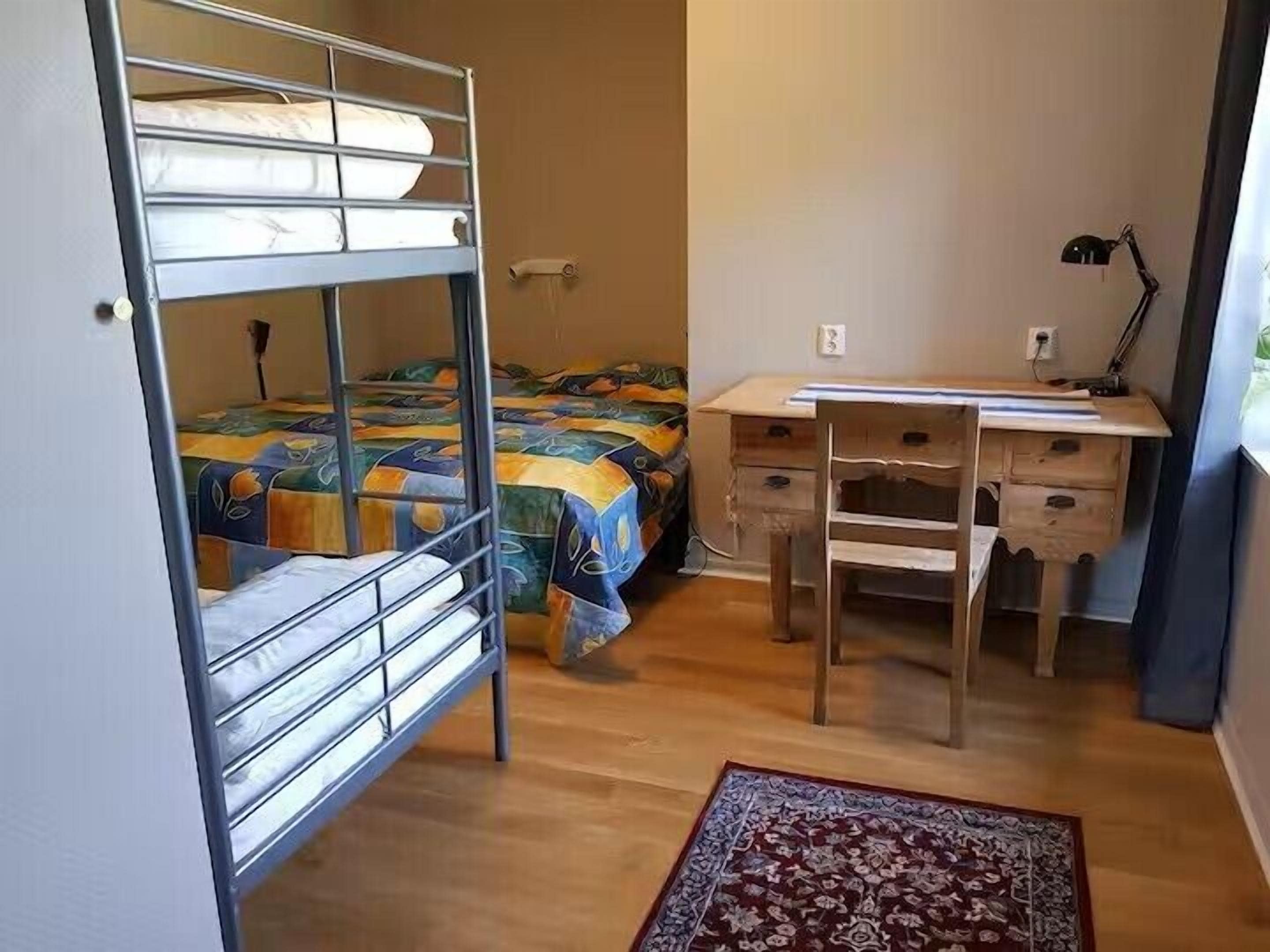 A dormitory room with bunk beds, a desk, a chair, a lamp, and a patterned rug.