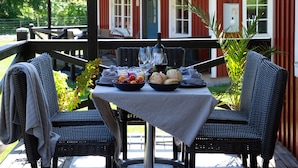 An outdoor patio with a table set for two, surrounded by wicker chairs, a covered area, and a red building in the background.