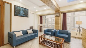 A modern hotel room with a blue sofa, a glass coffee table, and large windows with curtains. - Golden Dolma (Darjeeling)
