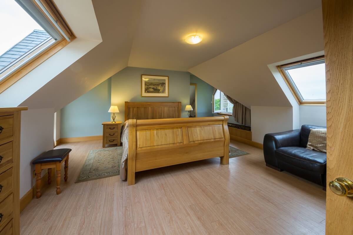 A bedroom with a sloped ceiling, a wooden bed, a black leather sofa, a wooden dresser, and a small table with lamps.