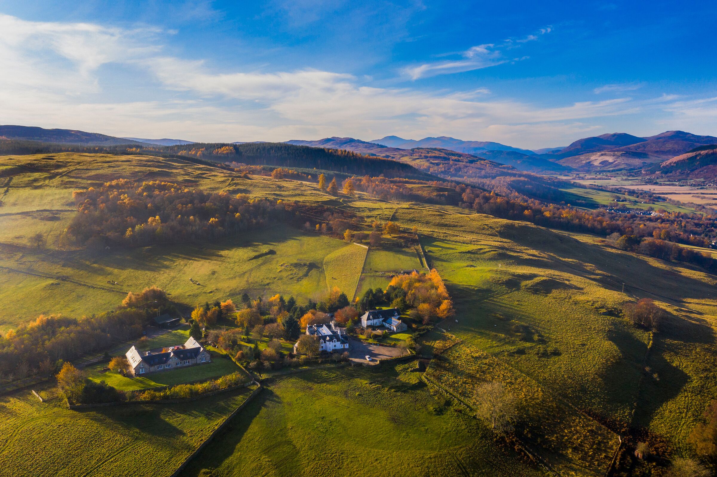 Vista aérea de un paisaje rural con colinas ondulantes, un pequeño conjunto de casas blancas rodeado de campos y montañas distantes bajo un cielo despejado.