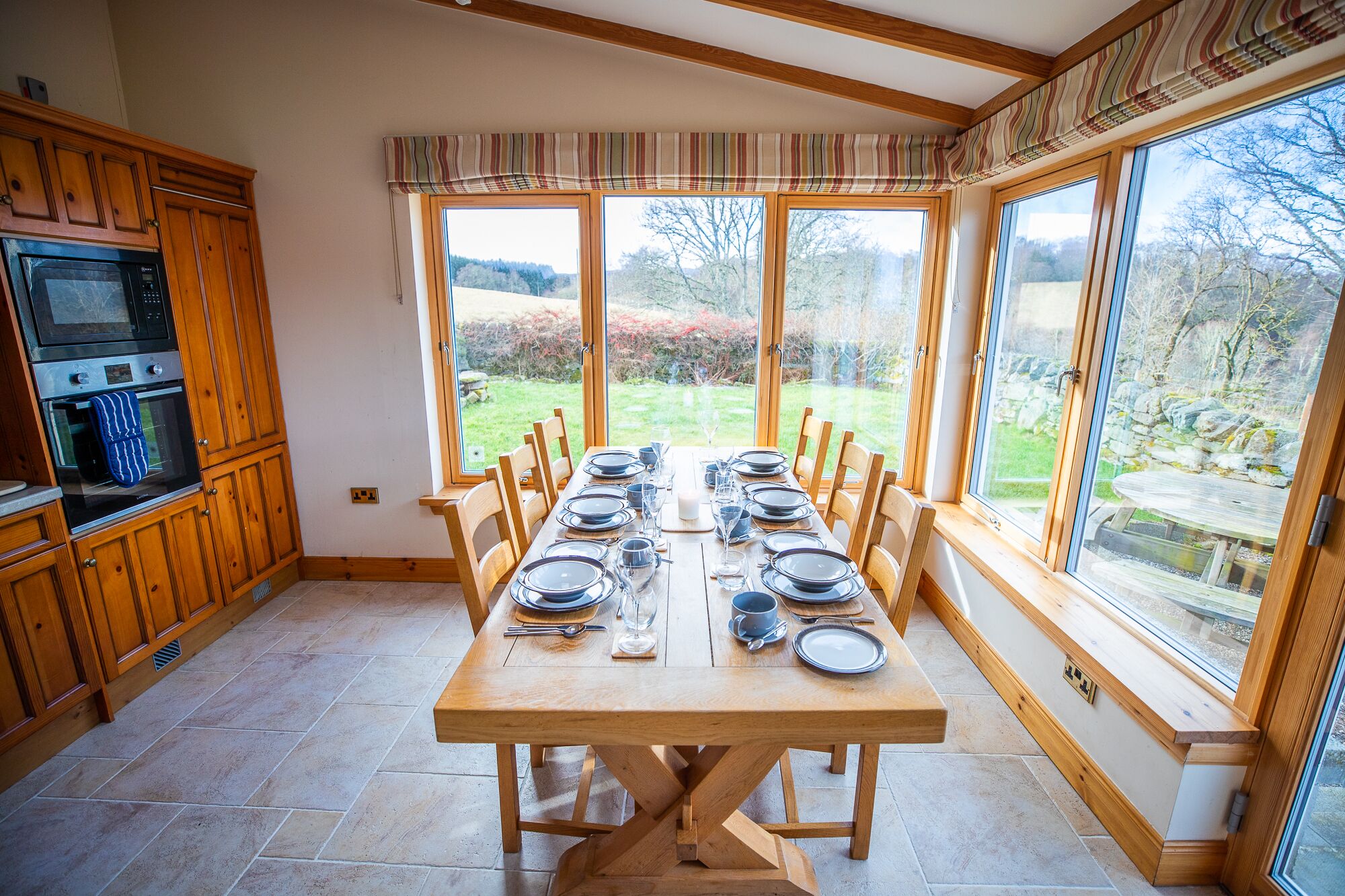 Comedor con mesa de madera dispuesta para seis personas, flanqueado por grandes ventanales que dan a un jardín.