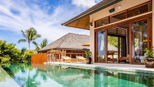 Une piscine aux eaux cristallines, dotée de chaises longues et d’une terrasse en bois, le tout entouré d’une végétation luxuriante et d’une maison moderne aux grandes fenêtres.