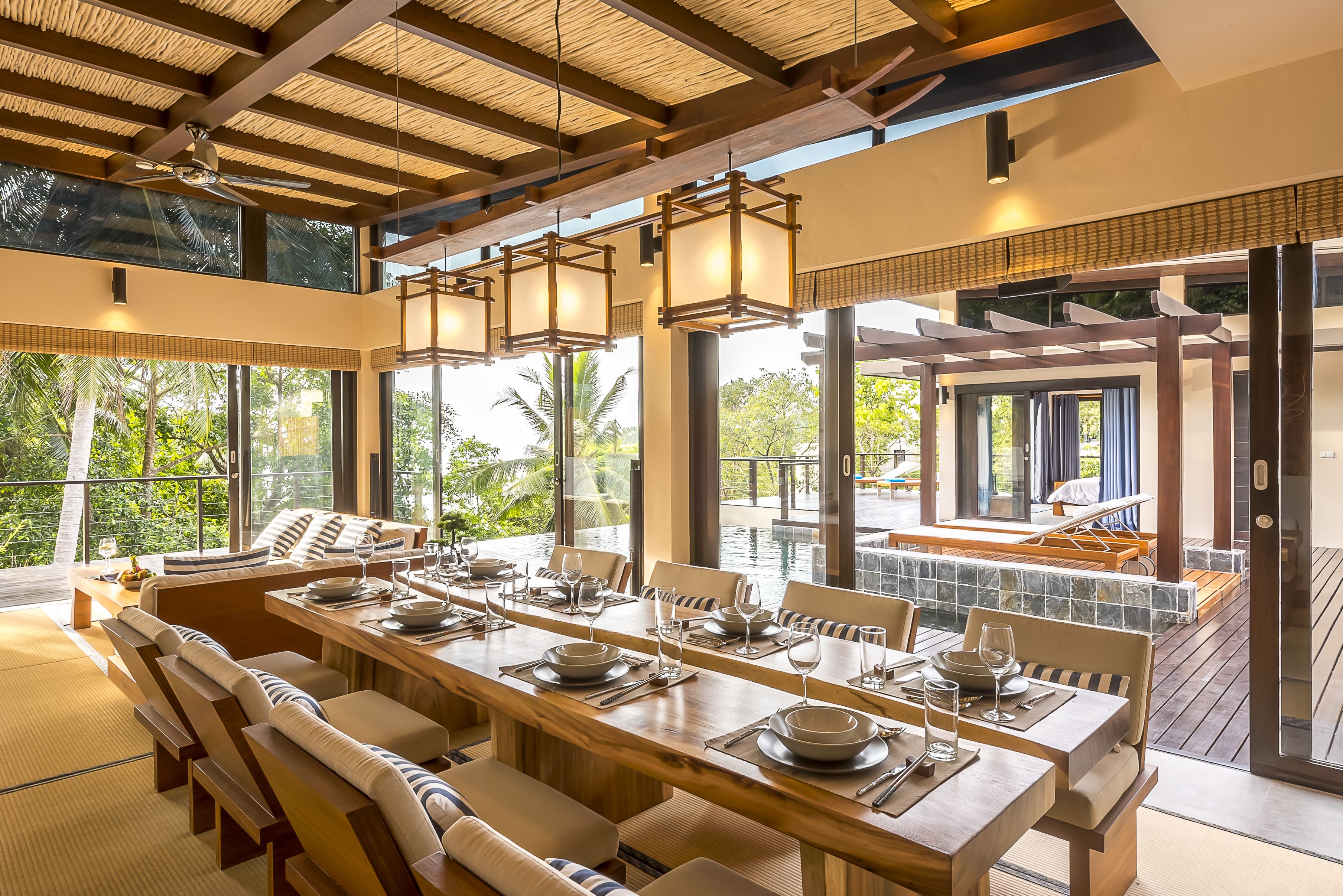 A spacious dining area with a long wooden table set for six, surrounded by cushioned seating. Large windows provide a view of the outdoors.