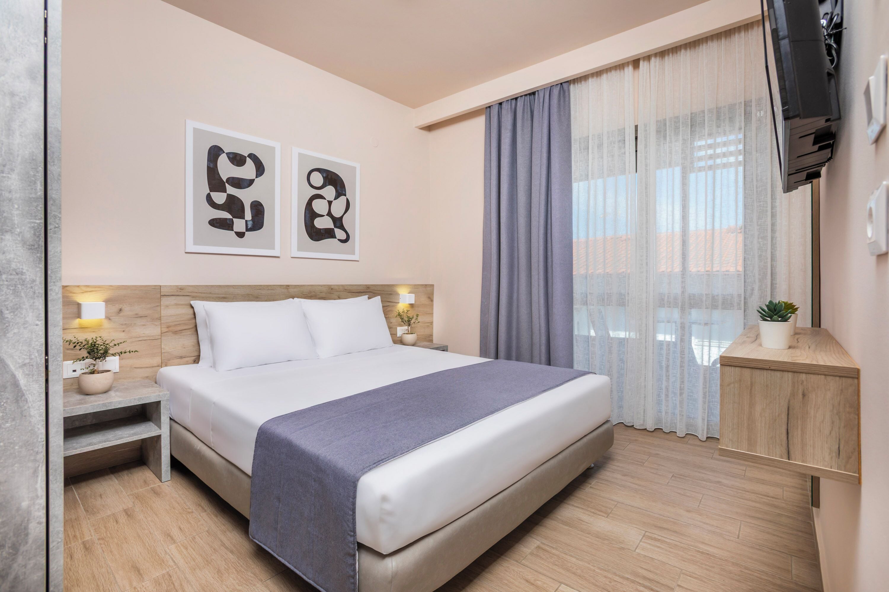 Habitación de hotel moderna con una cama grande, cabecera de madera, mesita de noche con lámpara y una pequeña planta en maceta.