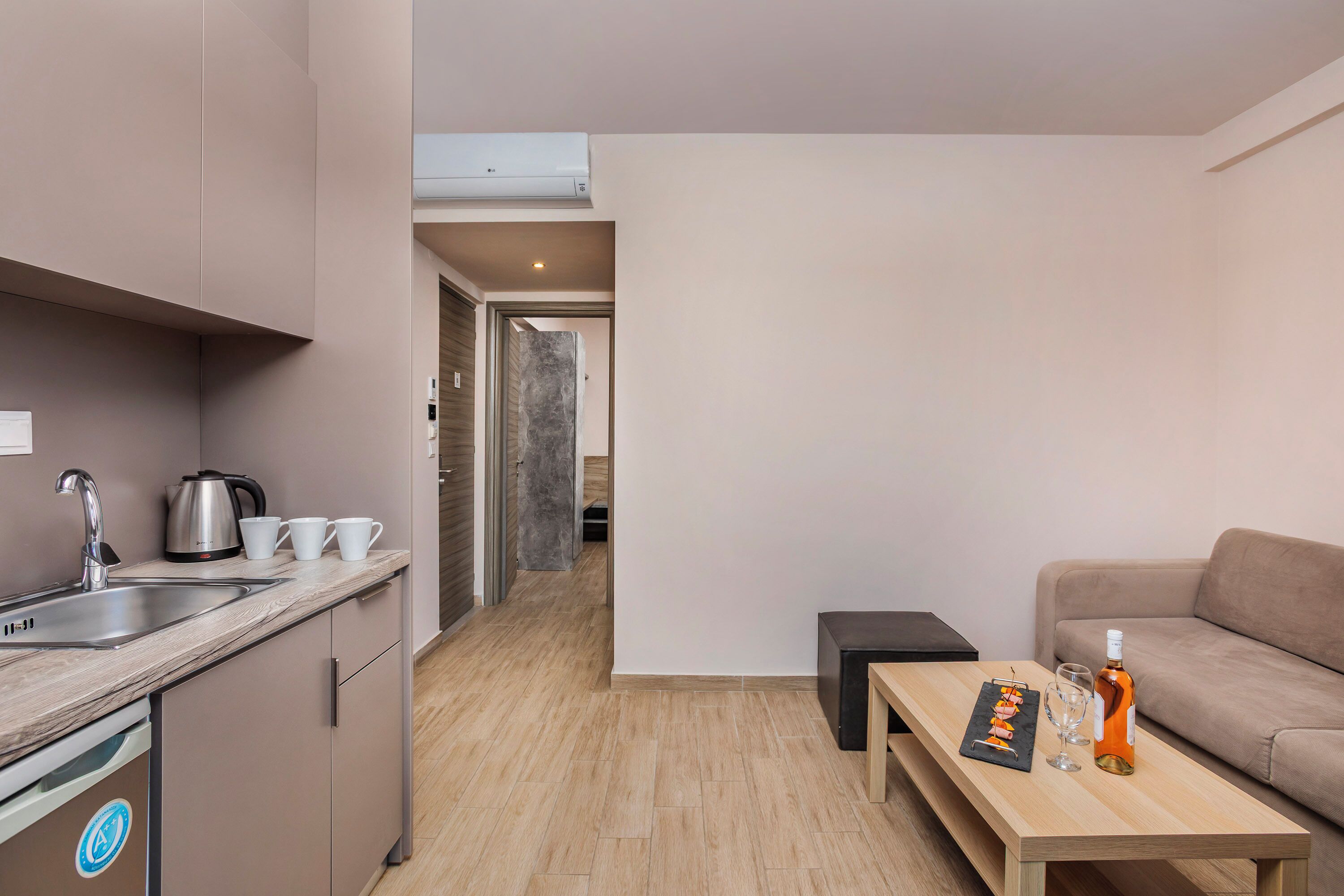 A modern kitchen with a sink, kettle, and cups, a wooden table with a bottle and glasses, and a sofa in a living area.