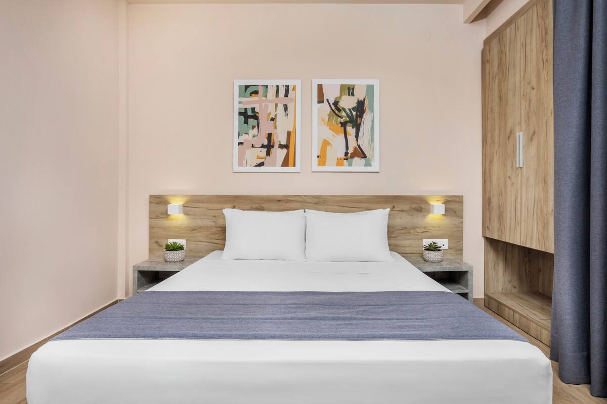 A modern bedroom with a wooden headboard, two bedside tables with lamps, a bed with white linens, and abstract art on the wall.