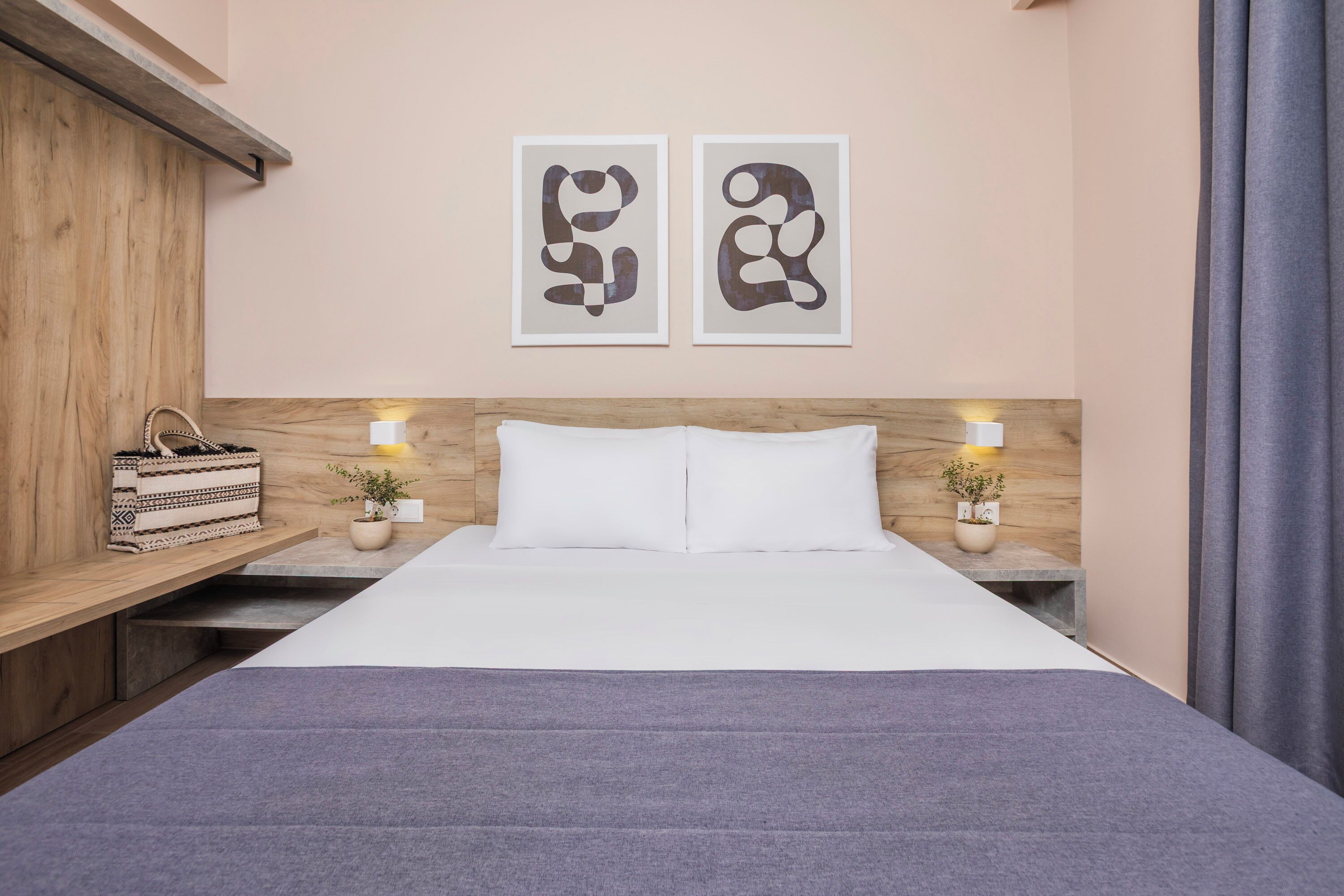 A neatly arranged hotel room with a wooden headboard, two bedside tables with lamps, a bed with white linens, and two abstract wall art pieces.