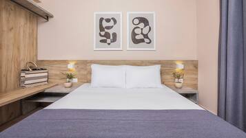 A neatly arranged hotel room with a wooden headboard, two bedside tables with lamps, a bed with white linens, and two abstract wall art pieces.