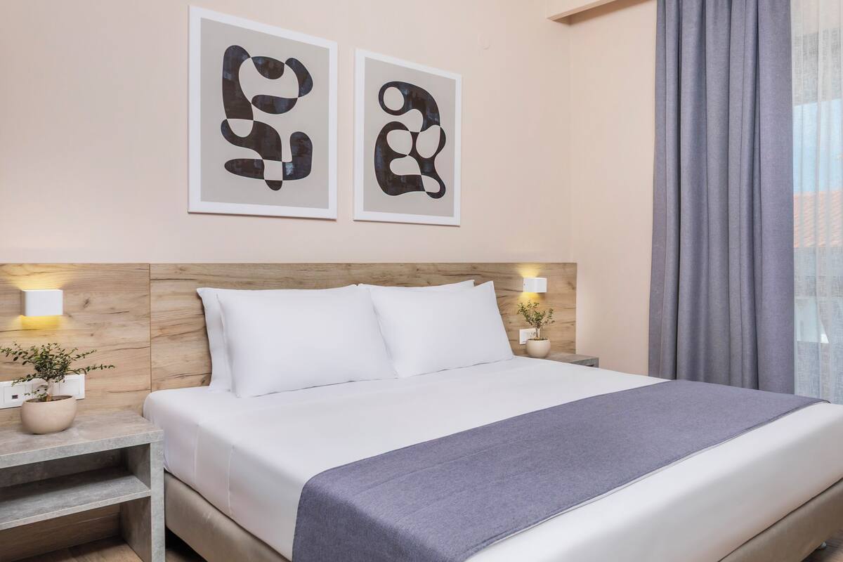 A neatly made bed with white linens and a gray throw blanket, a wooden headboard, two wall-mounted abstract art pieces, and a nightstand with a plant and a lamp.