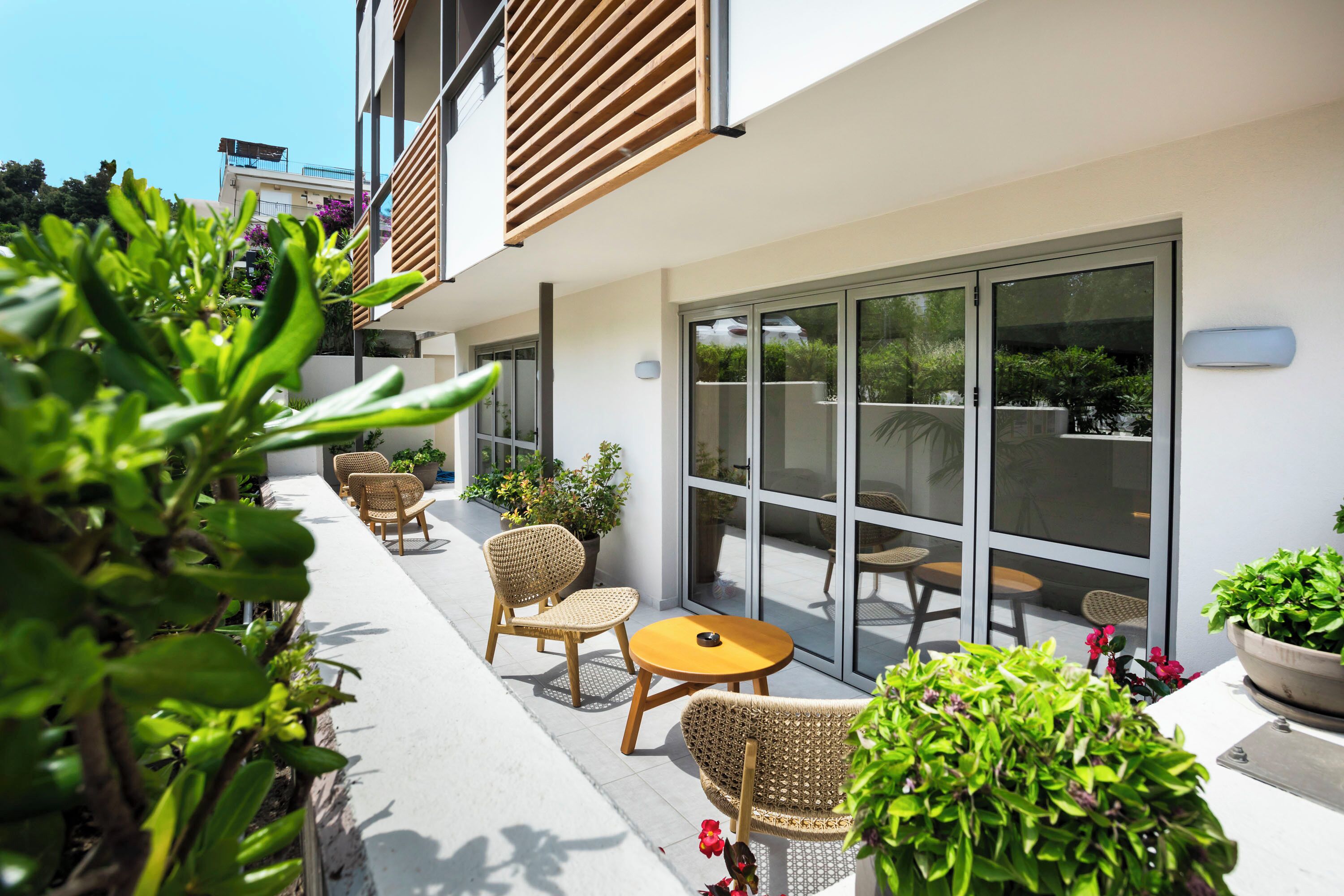Un espace extérieur aménagé avec des chaises en osier et une table ronde, entouré de plantes en pot et doté d’une porte vitrée donnant sur un patio couvert.